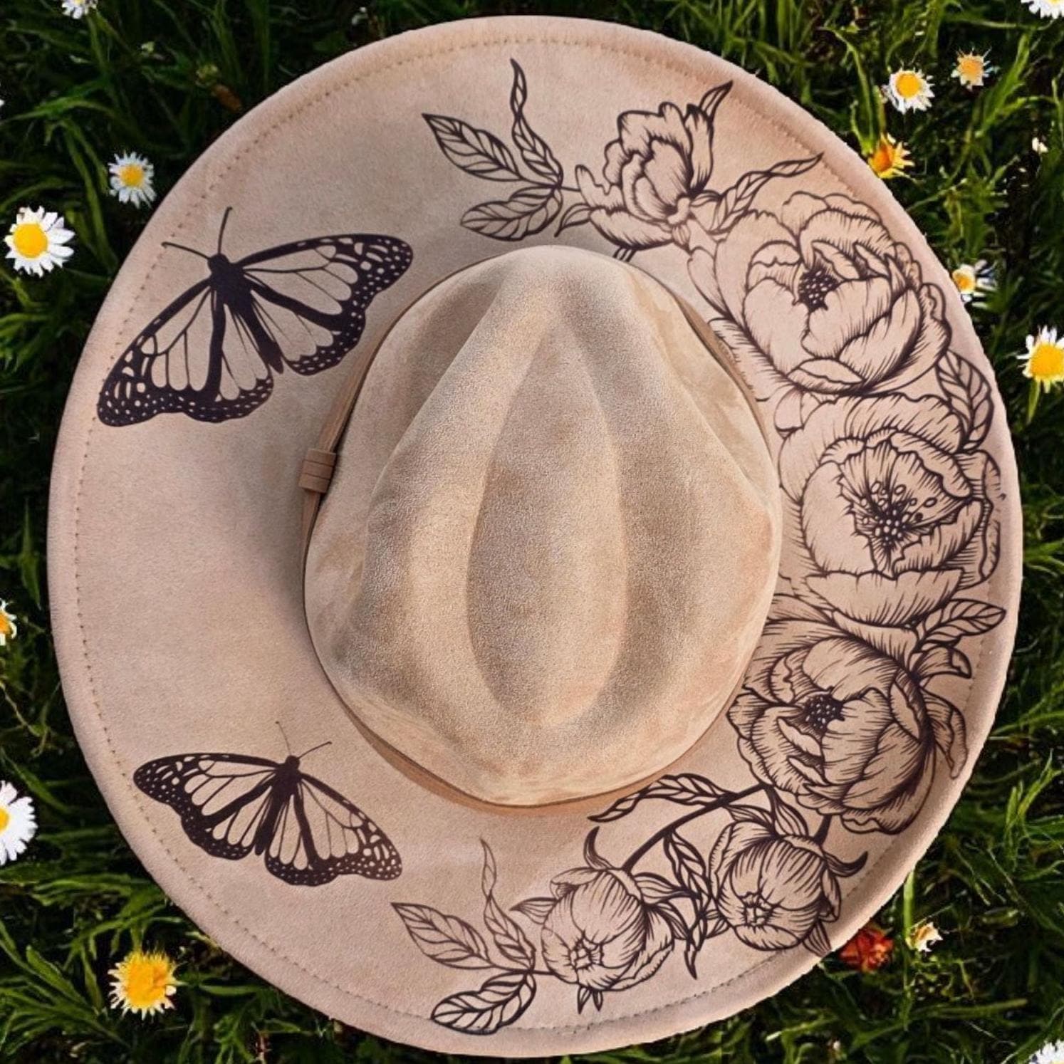 Laser engraved butterfly and floral design on tan vegan suede wide brim hat photographed in a daisy field
