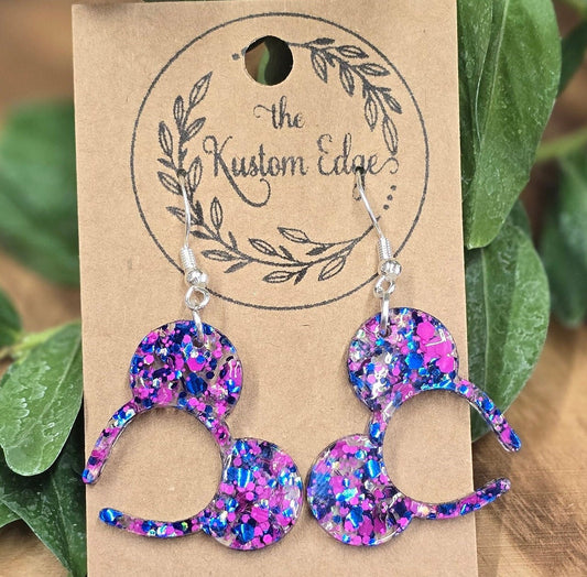 May include: A pair of silver dangle earrings with blue, pink, and silver glitter resin Mickey Mouse ears. The earrings are on a brown card with the text 'the Kustom Edge'.