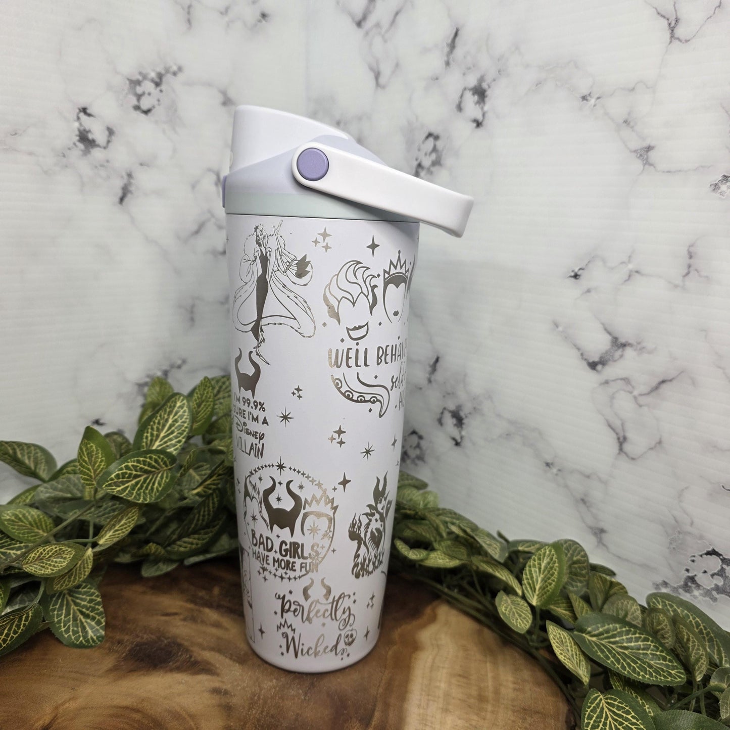 A white ceramic mug with various printed designs and text on it, placed on a wooden surface surrounded by green leaves.