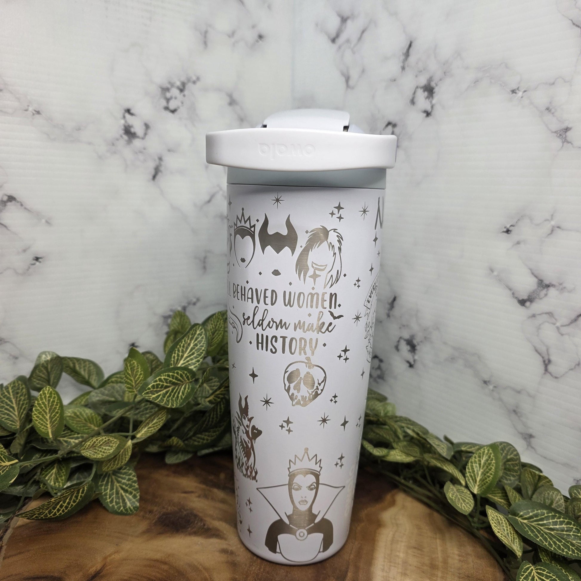 A white ceramic mug with a metallic design and text printed on it, placed on a wooden surface with green leaves.