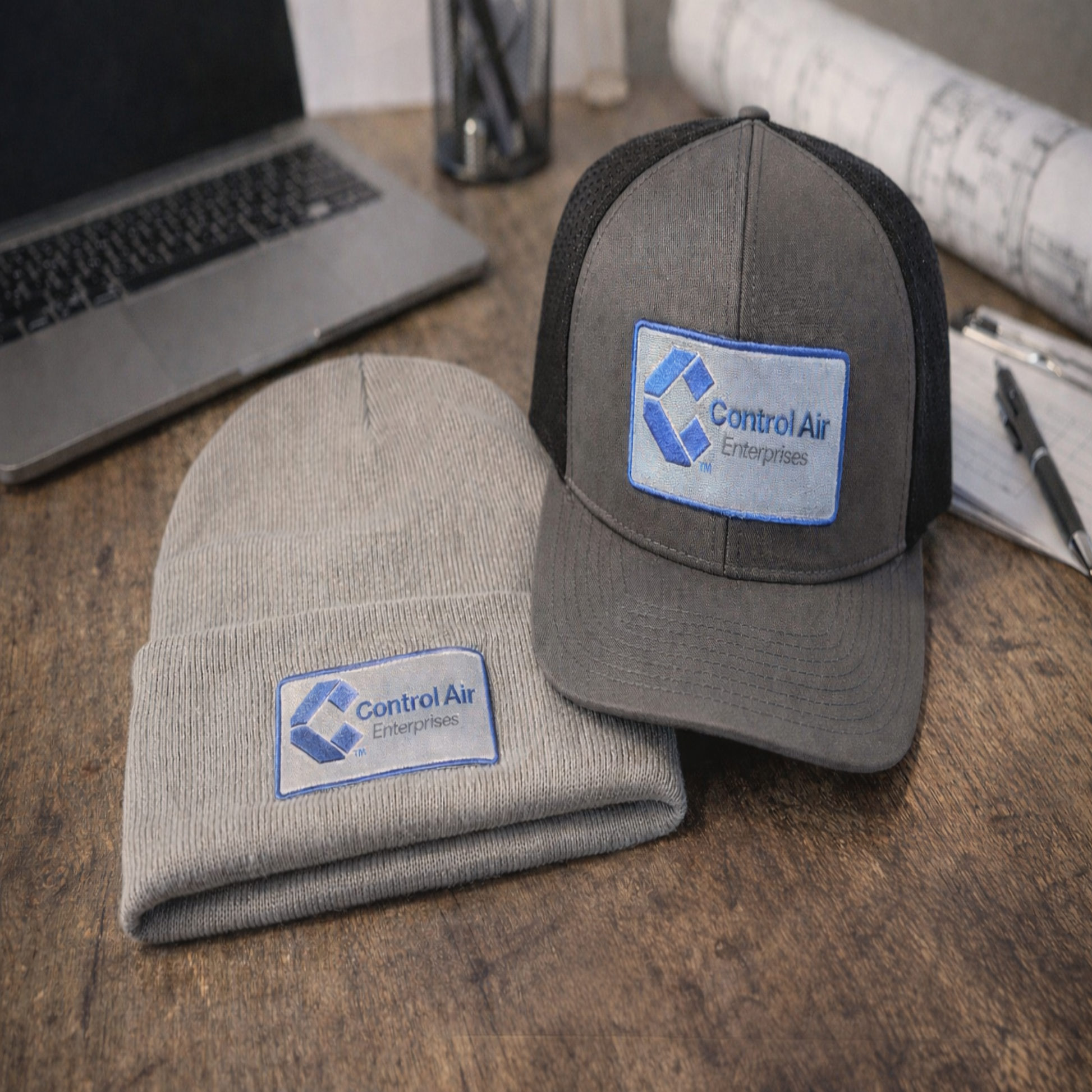 Gray beanie and cap with Control Air Enterprises logo on a wooden surface.