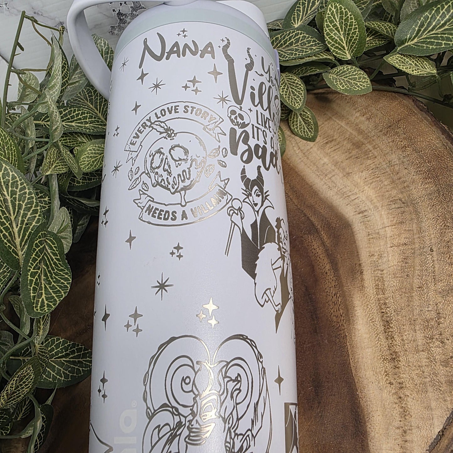 a white mug with various designs and text printed on it, placed on a wooden surface surrounded by green leaves.