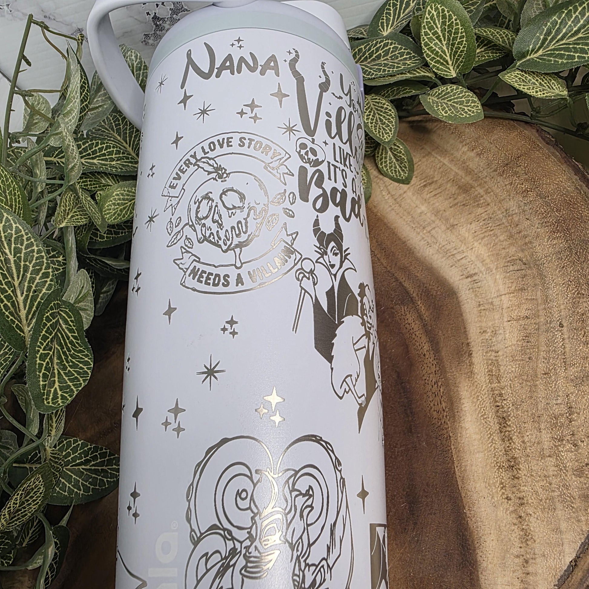 a white mug with various designs and text printed on it, placed on a wooden surface surrounded by green leaves.