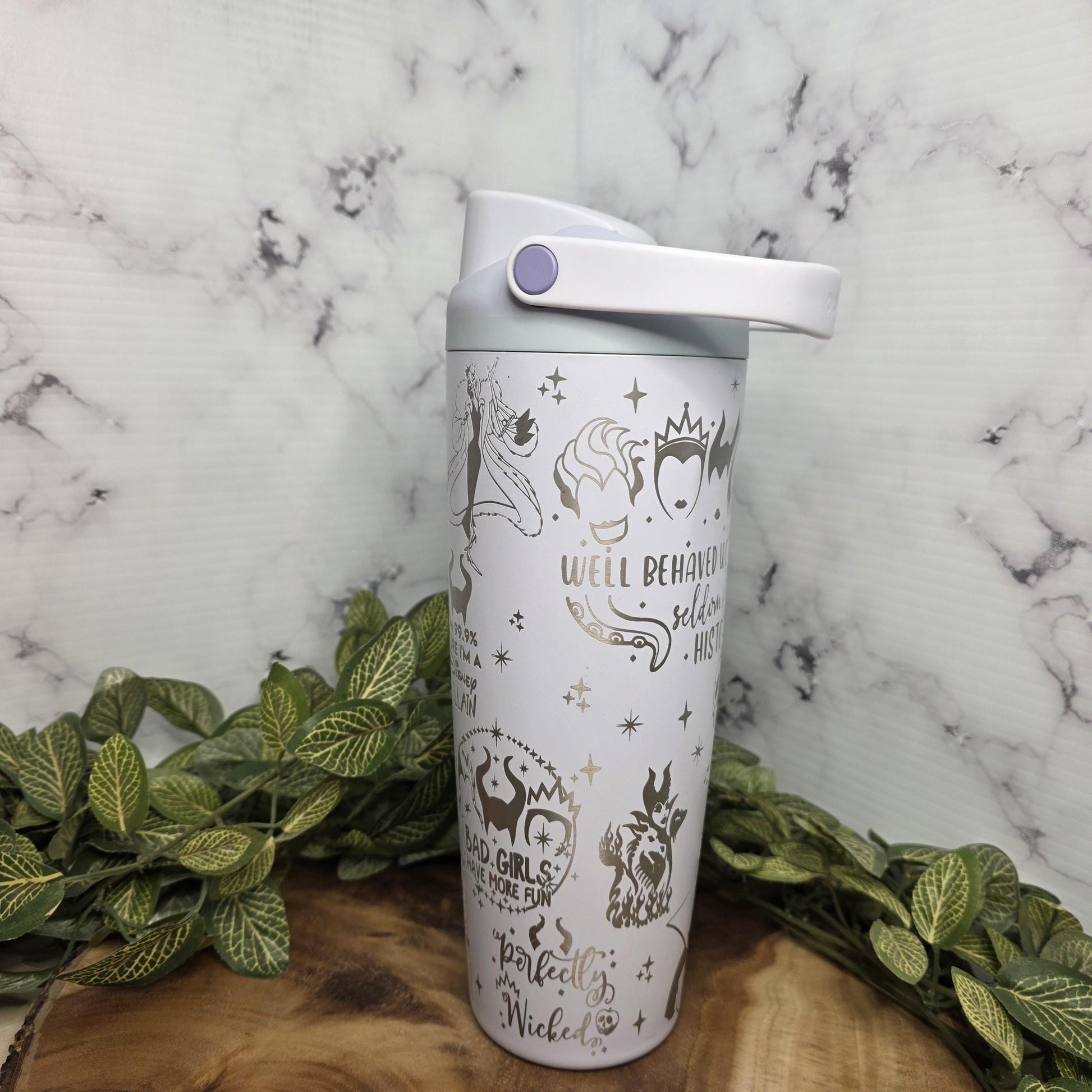 A white ceramic mug with various printed designs and text on it, placed on a marble surface with greenery.